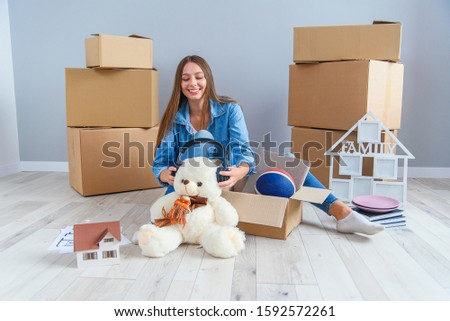 Beautiful smiling girl puts on the headphones on the teddy bear toy and sitting on the floor in the empty new apartment.