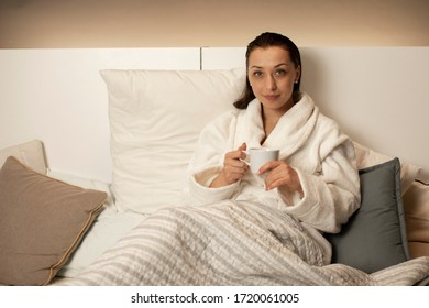 Beautiful Smiling Girl With Wet Hair After Shower Relaxing At Home Sitting In Bed Drinking Herbal Tea In Bathrobe. Young Brunette Woman Resting Before Bedtime. Chilling, Calmness, Harmony Concept