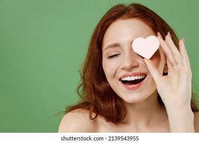 Beautiful Smiling Half Naked Topless Redhead Woman 20s Do Nude Make Up Cover Eye With Heart Shape Sponge Isolated On Pastel Green Color Background. Skin Care Healthcare Cosmetic Procedures Concept.