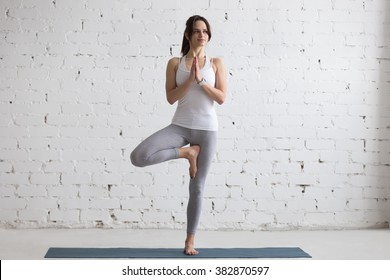 Beautiful Smiling Happy Young Woman Working Out Indoors, Doing Yoga Exercise On Blue Mat, Standing In Vrksasana Posture, Tree Pose, Full Length