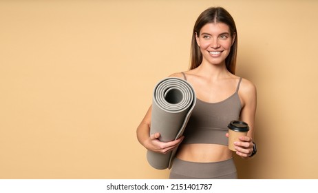 Beautiful Smiling Healthy Female Fitness Trainer In Stylish Brown Crop Top And Leggings Holding Rolled Mat And Plastic Cup Of Coffee After Workout Classes Over Peach Color Background