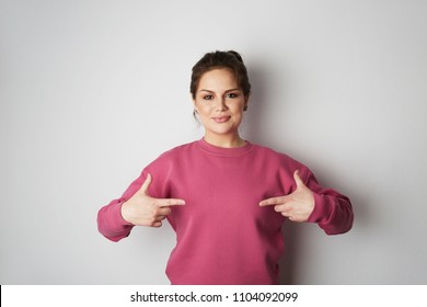 Beautiful smiling hipster girl wearing pink hoody and pointing finger with space for your logo or design. Mock-up of pink hoody on white empty wall in the background. Copy paste text space