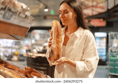 Hermosa mujer sonriente de Latina disfrutando del aroma de una baguette recién horneada mientras compra en una panadería de supermercado, rodeada de varios productos de pan y pasteles. Concepto alimentario