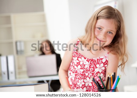 Beautiful smiling little blonde girl in a pretty floral summer frock inside in a classroom or home office with an adult woman working on a laptop and supervising her visible in the background