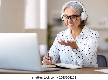 Beautiful smiling mature woman in headphones during online meeting on laptop with colleagues at home, senior female participating in virtual class, learning foreign language remotely at home