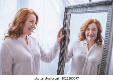 beautiful smiling mature woman standing near mirror and looking at reflection