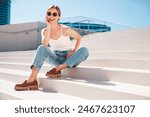 Beautiful smiling model in sunglasses. Female dressed in summer white T-clothes. Posing near white wall in the street. Funny and positive woman having fun outdoors. Sits at stairs, in sunglasses