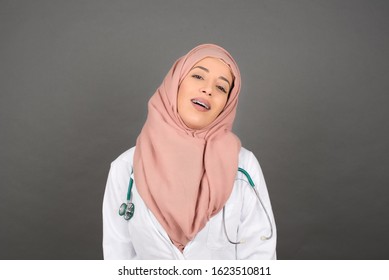 Beautiful Smiling Muslim Doctor Female With Broad Smile, Shows White Teeth, Wears Medical Uniform, Stands Over Gray Studio Wall Rejoices Having Day Off.