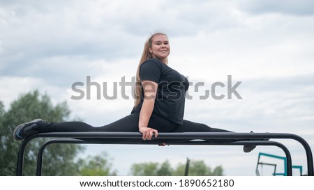 A beautiful smiling overweight young woman stretches for split on uneven bars outdoors. Fat girl gymnast doing fitness on the sports field. Sport outside on a warm summer day.