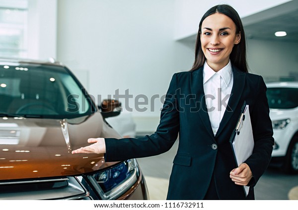 Beautiful smiling\
saleswoman in full suit in dealership on cars background with\
documents in tablet in\
hands