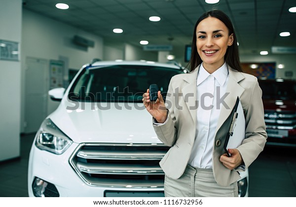 Beautiful smiling\
saleswoman in full suit in dealership on cars background with car\
keys and documents in\
hands
