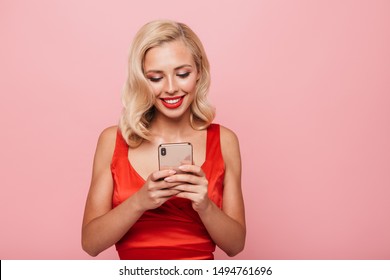 Beautiful Smiling Sexual Young Blonde Woman Wearing Red Cocktail Dress Standing Isolated Over Pink Background, Using Mobile Phone