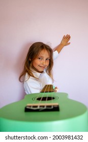 Beautiful Smiling Tanned Caucasian Child Kid Girl In A White Shirt Holds A Bright Ukulele Guitar In Her Hand At A Music Lesson At A Music School On A Uniform Background