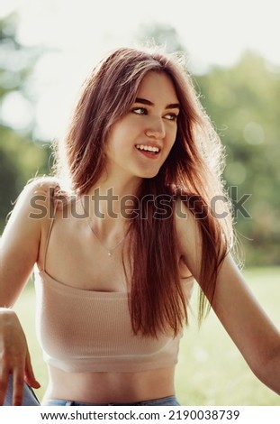 Beautiful smiling teen woman looking happy and making funny mug and showing the tongue sitting on the green glass outdoors summer green trees background. Closeup portrait in sunny day