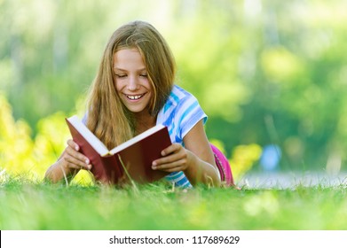 Beautiful Smiling Teenage Girl In Blue Blouse Lying On Grass And Read Book, Against Green Of Summer Park.