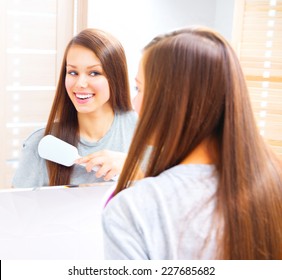 Beautiful smiling teenage girl is combing her long silky hair after getting up in the morning. Young attractive happy woman is brushing her brown hair enjoying them in the mirror and admiring herself 