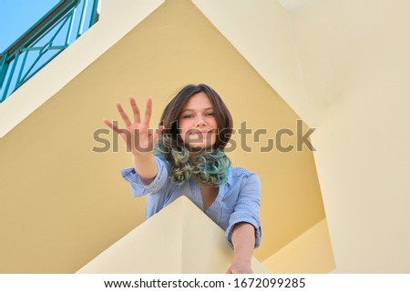 Beautiful smiling teenage girl looking at camera waving greeting, looking down standing on balcony, sunny summer day