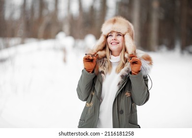 Beautiful Smiling woman admiring Winter nature in forest, wearing fur khaki jacket, ear flaps hat and leather gloves, cozy portrait at snow background