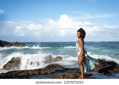 Beautiful smiling woman with black power hair wearing light green outfit with white bikini standing on the rocks of a beach. Blue sky and clouds in the background.  - Powered by Shutterstock