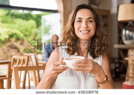 Beautiful smiling woman drinking coffee at cafe. Portrait of mature woman in a cafeteria drinking hot cappuccino and looking at camera. Pretty woman with cup of coffee.