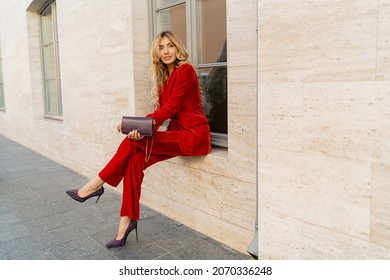 Beautiful Smiling Woman  In  Elegant Red Velvet Suit Holding Purse And  Posin Outdoor In Old European City.    Blond Wavy Hairs, Perfet Skin, Full Lips. 