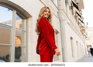 Beautiful Smiling Woman  In  Elegant Red Velvet Suit Holding Purse And  Posin Outdoor In Old European City.    Blond Wavy Hairs, Perfet Skin, Full Lips. 