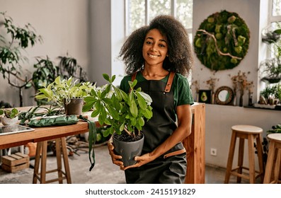 Beautiful smiling woman florist gardener professional in green t-shirt and apron holding pot with avocado plant and smiling looking at camera. Cultivation and sale of plants. - Powered by Shutterstock