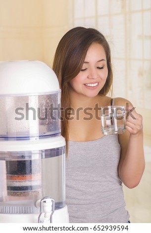 Beautiful smiling woman holding a glass of water behind of a filter system of water purifier on a kitchen background