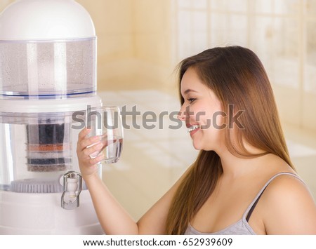 Beautiful smiling woman holding a glass of water, with a filter system of water purifier on a kitchen background
