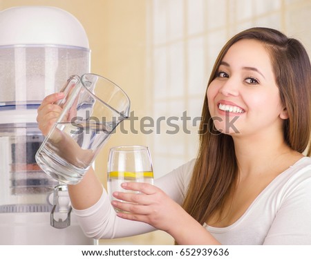 Beautiful smiling woman holding a glass of water in one hand and a pitcher of water in her other hand, with a filter system of water purifier on a kitchen background