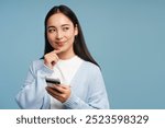 Beautiful, smiling woman holding smartphone is thinking about something, looking away isolated on blue background. Online shopping concept