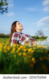Beautiful smiling woman in plaid shirt and jeans lying on a grass outdoor. Beautiful woman lying down in the meadow of flowers. Pretty girl relaxing outdoors on spring grass
