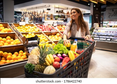 Beautiful smiling woman pushing shopping cart and taking fruits off the shelves in supermarket. Buying food at grocery store.