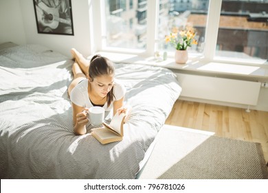 Beautiful Smiling Woman Reading Book And Enjoying Warm Coffee In Sunny  Apartment.Relaxation In Bed.Sunny Light Warm Home.Hygge Scandinavian Concept