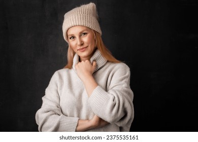 Beautiful smiling woman standing at isolated dark background. Blond haired woman wearing knit hat and turtleneck sweater. Copy space.  - Powered by Shutterstock