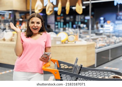 Beautiful Smiling Woman Standing In A Large Grocery Store Near A Grocery Cart, Holding A Mobile Phone And Looking At The Camera In Surprise. The Woman Is Shocked In The Store