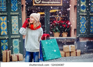 Beautiful  smiling woman at the street with shopping bags talking on mobile phone.
Attractive girl  dressed in warm white  hat, red scarf and mittens. Magic snowfall effect.
