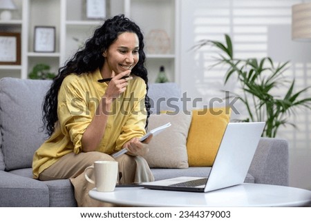 Beautiful smiling woman studying online remotely sitting on couch in living room inside house, Hispanic woman using laptop to watch course video, writing thesis in notebook.