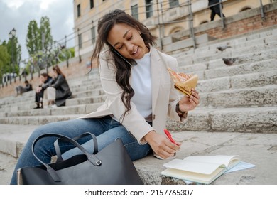 Beautiful smiling woman talking on mobile phone, taking notes, eating slice of tasty pizza on the street. Stylish businesswoman working outdoors , multitasking concept 