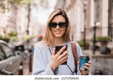 Beautiful smiling woman walking on street with coffee takeaway, chatting on mobile phone. Girl talking on smartphone on her way to meeting spot, sending message, wear blue sweater and black glasses. - Powered by Shutterstock