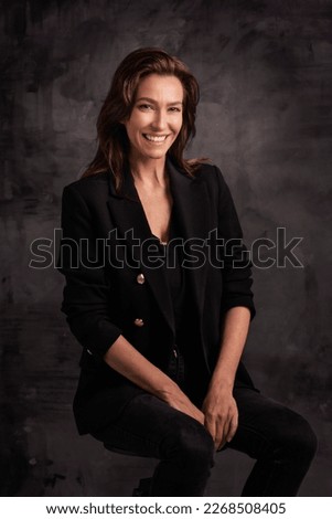 Beautiful smiling woman with white teeth sitting at isolated dark grey background. Happy woman wearing black blazer. Copy space. 