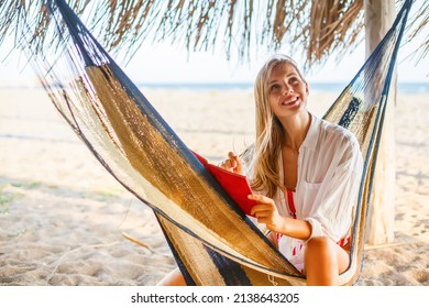 Beautiful smiling woman writing something in her red planner sitting in a hammock on a beach. Life or job planning in comfortable environment.