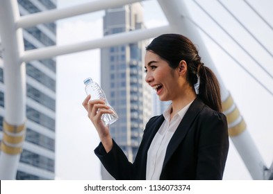beautiful smiling young asian business woman wearing modern black suit holding a bottle of natural water for refresh yourself with modern city background, lifestyle, healthy, summer concept