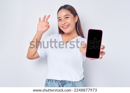 Beautiful smiling young Asian girl wearing casual white t-shirt holding mobile phone with blank screen, gesturing okay sign isolated on white background. People lifestyle concept
