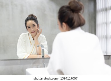 Beautiful smiling young Asian woman in bathrobe applying moisturizer cream on her pretty face and looking to mirror at bathroom, touching face with hands. Natural skin care and people Concept.
