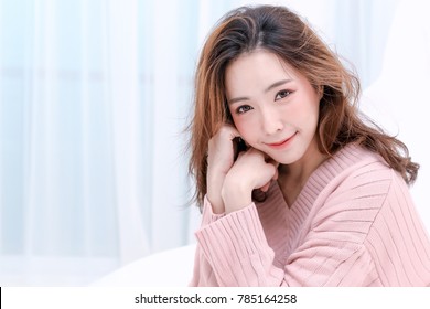 Beautiful Smiling Young Asian Woman with Clean, Fresh, Glow, and pefect Skin in pink sweater, winter concept. Happy, healthy, cheerful, relaxing girl in white living room.