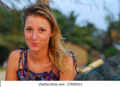 Beautiful, smiling young blond girl sitting on tropical beach with fishing nets in background.