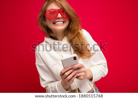 Beautiful smiling young blonde woman wearing stylish white hoodie and funny colorful glasses standing isolated over colourful background holding and using mobile phone communicating online on the