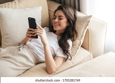 Beautiful smiling young brunette woman relaxing on a couch at home, using mobile phone