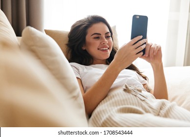 Beautiful Smiling Young Brunette Woman Relaxing On A Couch At Home, Using Mobile Phone